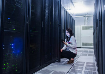 A female data center technician is kneeling next to a server rack, checking on data results on her laptop.