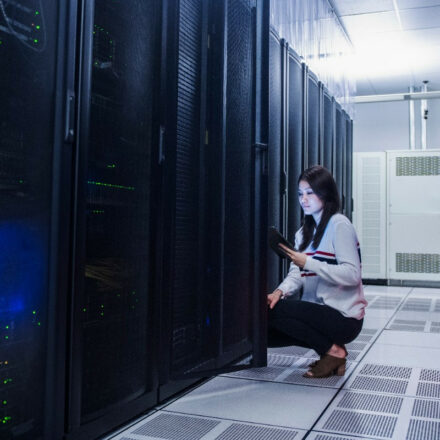 A female data center technician is kneeling next to a server rack, checking on data results on her laptop.