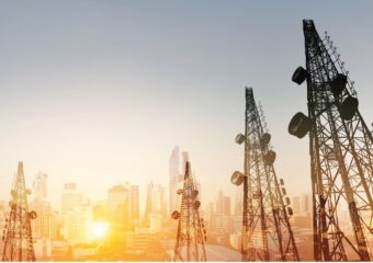Telecommunications towers in the foreground as the sun rises against a cityscape in the background.