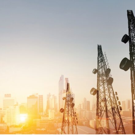 Telecommunications towers in the foreground as the sun rises against a cityscape in the background.