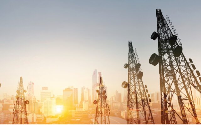 Telecommunications towers in the foreground as the sun rises against a cityscape in the background.