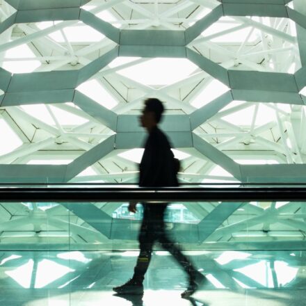 Defocused image of a man walking in a modern design corridor, with a hexagonal design wall or building section appears in the background.
