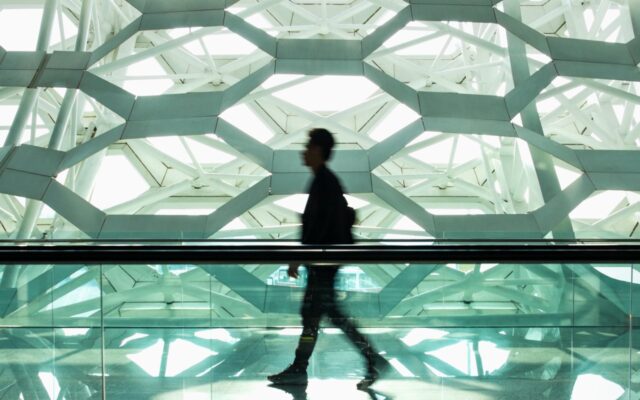 Defocused image of a man walking in a modern design corridor, with a hexagonal design wall or building section appears in the background.