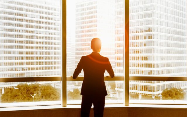 View from behind of a businessman looking out office windows at nearby buildings and sunlight emerging between buildings.