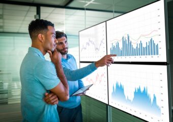 Two male colleagues in an office looking at reviewing various reports projected on to four Dell UltraSharp U2718Q monitors.