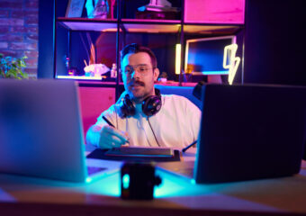 Young attractive man podcaster in glasses explaining concepts with stylus on tablet, in modern home-studio in neon light. Concept of youth people with social media and smart working, lifestyle, online